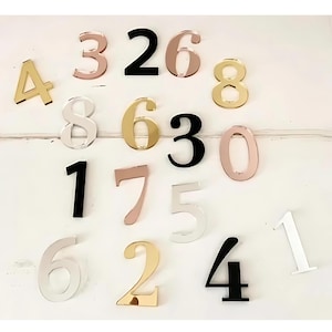 May include: Assortment of acrylic numbers in various colors, including gold, silver, black, white, and rose gold. The numbers are arranged in a random pattern on a white surface.