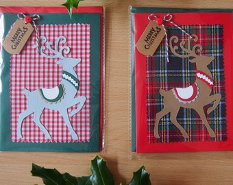 Puede incluir: Dos tarjetas de Navidad con recortes de renos. Una tarjeta tiene un fondo a cuadros rojos y blancos, la otra un fondo a cuadros rojos y negros. Cada tarjeta tiene una etiqueta que dice "FELIZ NAVIDAD". Las tarjetas están en fundas de plástico.