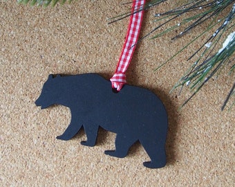 May include: A black bear ornament with a red and white checkered ribbon for hanging. The ornament is hanging in front of a sprig of evergreen and red berries, suggesting a holiday theme.