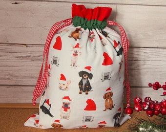 May include: A white drawstring gift bag featuring various dog breeds wearing Santa hats. The bag has a red and green ruffled top and is tied with red and white checkered ribbon. A festive holiday item.