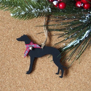 May include: A black wooden dog ornament with a red and blue plaid scarf and a small silver bell. The ornament hangs from a twine string. The background includes a pine branch with red berries and a pine cone.