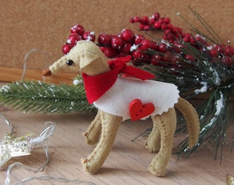 Pode incluir: Um enfeite de cão de feltro vestindo um suéter branco com um botão de coração vermelho e uma echarpe vermelha. O enfeite é cercado por um ramo de pinheiro nevado e luzes de Natal em forma de estrela prateada.
