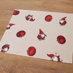 May include: A white placemat with a repeating pattern of red apples and gnomes. The gnomes are wearing red hats and have white beards.