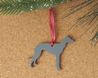 Pode incluir: Um ornamento de madeira cinza em forma de cão galgo, pendurado em uma fita xadrez vermelha e branca. O ornamento é suspenso de um galho de pinheiro verde com neve artificial.