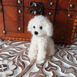 May include: A small, fluffy white dog figurine sits on a carved wooden surface in front of a miniature wooden chest. The chest is brown with metal accents and leather straps. The dog has black eyes and a black nose.