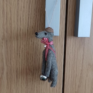 May include: A gray felt dog ornament with a brown hat and a red and white checkered bow. The dog has a silver bell around its neck and is hanging from a light gray rectangular tag. The ornament is hanging on a wooden cabinet.
