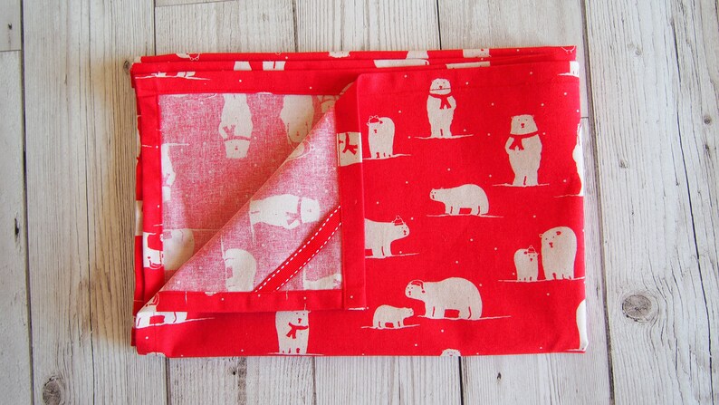 May include: Folded red fabric with a repeating pattern of white polar bears wearing scarves and hats. The fabric is folded to reveal a contrasting white interior with a red ribbon detail. The overall theme is winter or holiday.