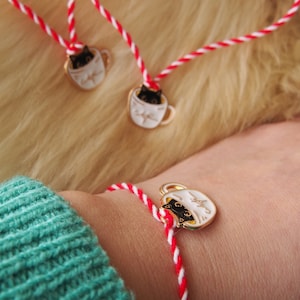 Op de afbeelding: Sieradenset met kat-in-mok bedels. De set bevat een ketting en armband, elk met een witte mok en een zwarte kat. De bedels zitten aan rood-witte gedraaide koorden.