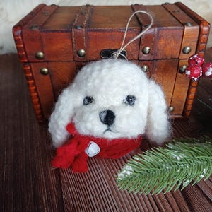 May include: A white, handcrafted dog ornament with black eyes and a black nose. The dog wears a red scarf with a small silver bell. The ornament is likely for Christmas or holiday decor.