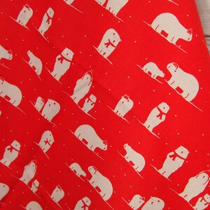 May include: Red fabric with a repeating pattern of white polar bears wearing red scarves. The bears are skiing down a diagonal slope, with small white dots scattered throughout the design. The fabric is against a light wood background.