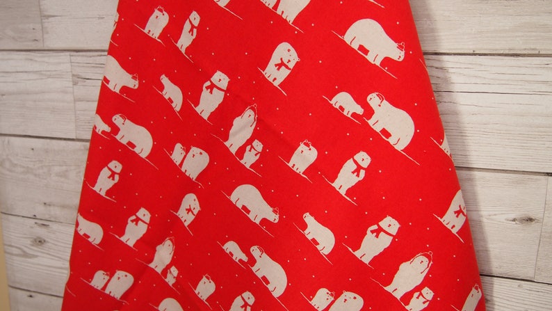 May include: Red fabric with a repeating pattern of white polar bears wearing red scarves. The bears are skiing down a diagonal slope, with small white dots scattered throughout the design. The fabric is against a light wood background.