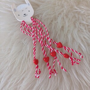 Op de afbeelding: Haaraccessoire met een konijnengezichtontwerp. Het heeft rood-witte gevlochten strengen, elk versierd met een kleine, hartvormige rode kraal. De achtergrond is een zacht, wit oppervlak dat op bont lijkt.