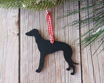 Pode incluir: Um enfeite de Natal com a silhueta preta de um cão, pendurado por uma fita xadrez vermelha e branca. O cão é de raça esguia, possivelmente um galgo. O enfeite está em frente a um fundo de madeira com ramos de pinheiro.
