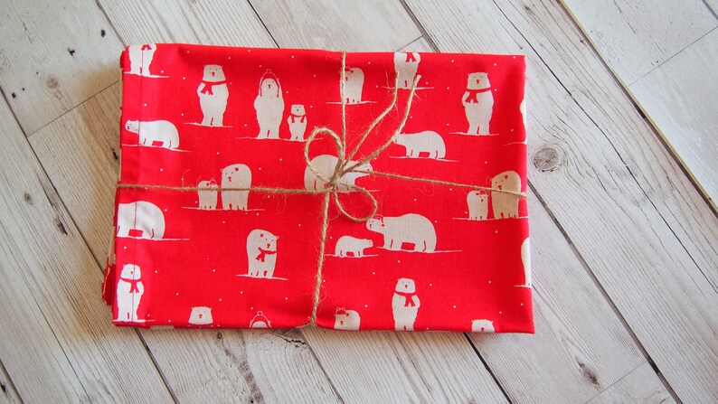 May include: A red fabric item wrapped with twine, featuring a repeating pattern of white polar bears wearing red scarves. The bears are in various poses, some with cubs. The fabric is folded and tied with natural twine.