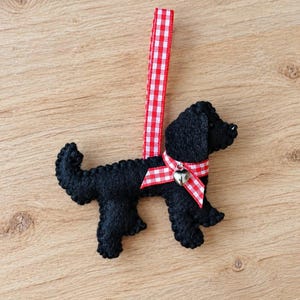 May include: A black felt dog ornament featuring visible stitched edges. The dog wears a red and white gingham ribbon collar with a small silver heart-shaped bell attached. A matching red and white gingham ribbon loop extends upwards for hanging.