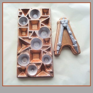 May include: A copper-coloured rectangular mould with geometric shapes and silver-coloured circular indentations. Beside it is a copper-coloured letter "A" with silver-coloured compass details. The items are likely for crafting or decorative purposes.