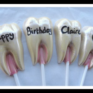Peut inclure: Quatre sucettes en forme de dents avec des bâtonnets blancs. Les sucettes sont décorées de glaçage doré et rose. Les sucettes sont décorées des mots "Happy", "Birthday", "Claire" et d'un cœur rose.