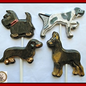 May include: Four chocolate dog lollipops on white sticks. The dogs are a black and white pointer, a black dachshund, a black and gold German Shepherd, and a black Scottish Terrier.