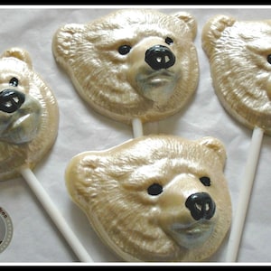 Puede incluir: Tres piruletas de chocolate blanco con forma de cabeza de oso sobre papel pergamino blanco. Las caras de los osos están detalladas con ojos y narices negros.