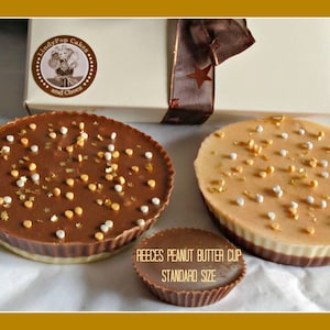 May include: Several large peanut butter cups with chocolate coating and sprinkles, alongside a smaller standard-sized cup labelled "Reeces Peanut Butter Cup Standard Size". A gift box with a ribbon and star is visible in the background. The treats are from LindyPop Cakes.