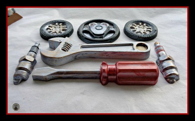 Puede incluir: Un surtido de elementos decorativos con tem&aacute;tica automotriz, que incluye dos buj&iacute;as, dos ruedas, un volante, una llave inglesa y un destornillador. Los elementos est&aacute;n pintados en plata met&aacute;lica, rojo y negro, dispuestos sobre una superficie blanca.