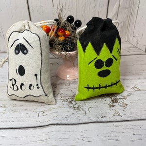 May include: Two Halloween-themed fabric bags with drawstring closures. One bag is white with a ghost face and the text "Boo!" The other bag is green with a Frankenstein face.