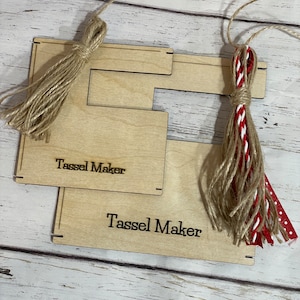 May include: Two wooden tassel makers with the words "Tassel Maker" engraved on them. One tassel maker has a natural jute tassel attached and the other has a red and white tassel attached.