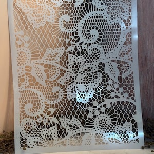 May include: A square, off-white stencil featuring a detailed lace pattern. The design includes various floral and geometric elements, creating an intricate and delicate aesthetic. The stencil is made of a flexible material, suitable for various craft projects.