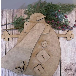May include: A wooden snowman decoration with a scarf, hat, and button details. The snowman has outstretched arms and a holly leaf design. The background includes evergreen branches and red berries, suggesting a holiday theme.