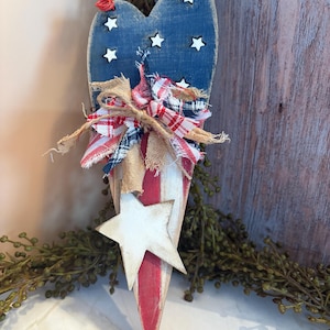 May include: A patriotic wooden heart-shaped decoration. The top is painted blue with white stars, transitioning to red and white stripes. A burlap and fabric bow adorns the center, and a white star is attached at the bottom. A red string is attached for hanging.