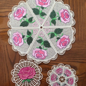 Könnte beinhalten: Drei weiße Häkeldeckchen mit gestickten rosa Rosen. Das größte Deckchen hat sechs Rosen, das mittlere Deckchen eine Rose und das kleinste Deckchen fünf Rosen.