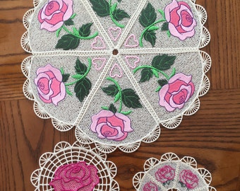 May include: Three white crocheted doilies with pink roses embroidered on them. The largest doily has six roses, the medium doily has one rose, and the smallest doily has five roses.