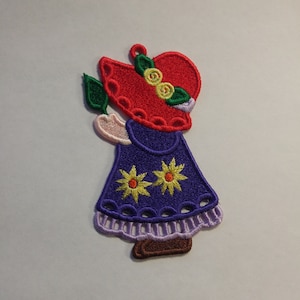 May include: Embroidered iron-on patch of a girl wearing a red hat with yellow flowers and a purple dress with yellow flowers.
