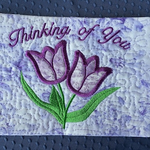 Könnte beinhalten: Eine weiße gesteppte Karte mit lilafarbener Stickerei von zwei Tulpen und dem Text "Thinking of You".