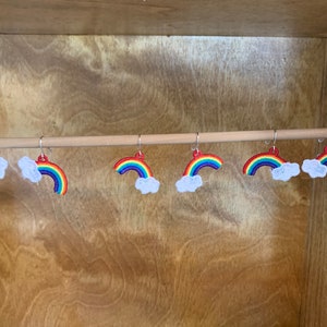May include: Six pairs of rainbow and cloud earrings hanging on a wooden rod. The earrings are made of colorful embroidery thread and feature a rainbow arch with a white cloud.