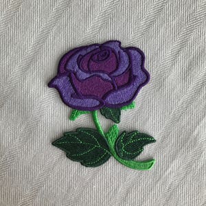 Puede incluir: Rosa bordada de color morado con hojas y tallo verdes. La rosa es una flor única con un diseño en capas. Las hojas están detalladas con un contorno cosido.