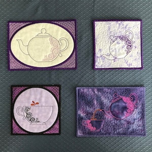 May include: Four purple and white quilted tea cosy squares with embroidered teapots and teacups. The teapots and teacups are outlined in black and have a swirl design.