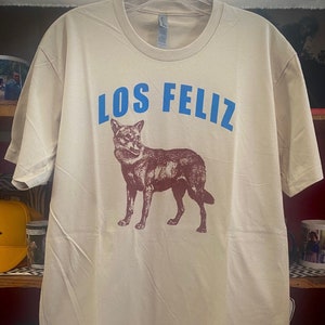 May include: A beige t-shirt with a brown wolf illustration and the text "LOS FELIZ" in blue lettering.