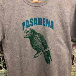 May include: A gray t-shirt with a teal blue graphic of a parrot perched on a branch. The text "PASADENA" is printed in teal blue above the parrot.
