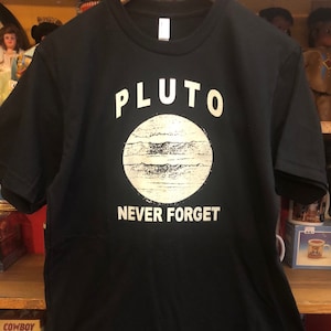 Könnte beinhalten: Schwarzes T-Shirt mit einer weißen Grafik von Pluto, einem Planeten mit Ringen, und dem Text "PLUTO NEVER FORGET".