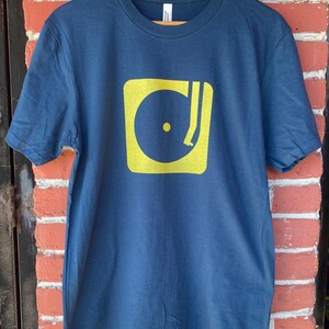 May include: A navy blue t-shirt featuring a yellow square graphic of a record player. The design includes a yellow record and tone arm. The shirt is hanging on a wooden hanger against a brick wall.