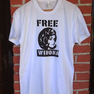 May include: White t-shirt with black text that reads "FREE WINONA" and a black and white illustration of a woman with a bouffant hairstyle.