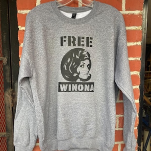 May include: A heather gray sweatshirt with the text "FREE WINONA" printed in black. The design includes a black and white image of a woman's face. The sweatshirt is hanging on a hanger against a brick wall.