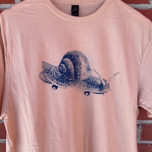 May include: A light pink t-shirt with a blue graphic of a snail skateboarding. The snail is wearing a shell and has a long, thin body.