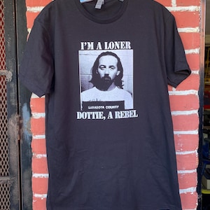 May include: Black t-shirt featuring the text "I'M A LONER" above a black and white mugshot of a man. Below the mugshot are the words "DOTTIE, A REBEL" and "SARASOTA COUNTY". The t-shirt is displayed on a hanger.