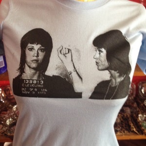 May include: Light blue t-shirt with a black and white image of a woman's mugshot. The mugshot includes the text "139813 CLEVELAND 32 5 8 126 NOV 3 1970".