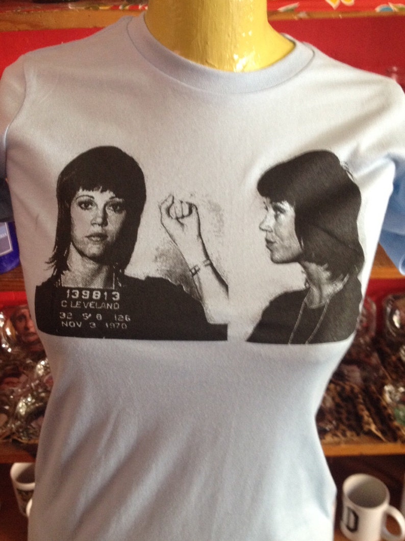May include: Light blue t-shirt with a black and white image of a woman's mugshot. The mugshot includes the text "139813 CLEVELAND 32 5 8 126 NOV 3 1970".