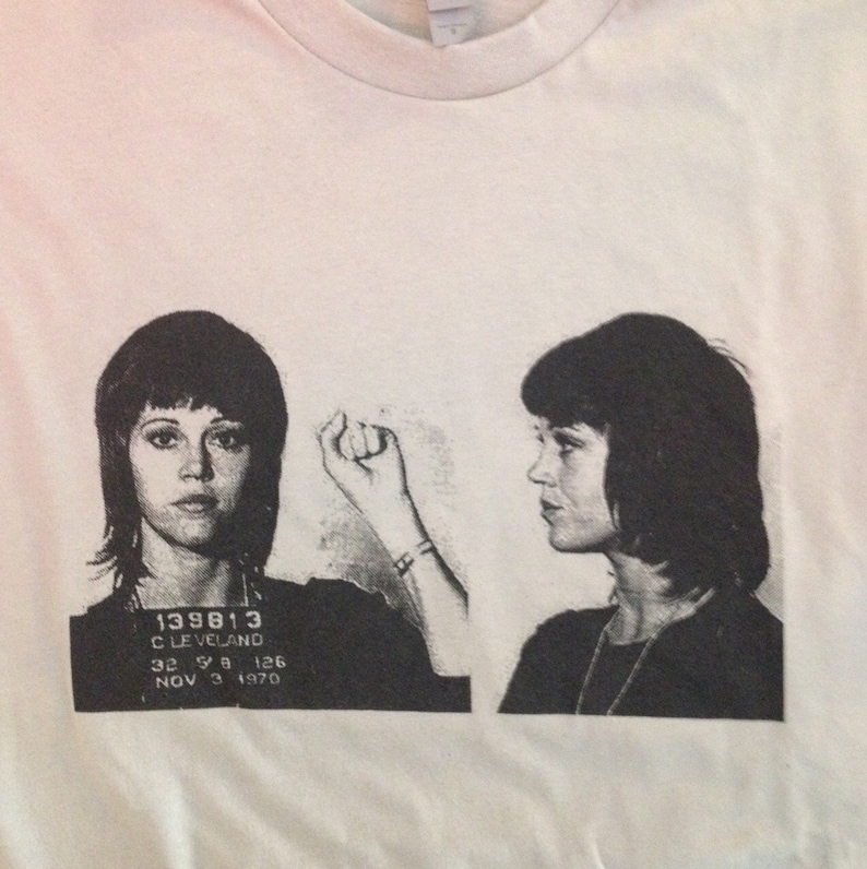 May include: Black and white mugshot style image of a woman with short dark hair. The image is printed on a white t-shirt. The text on the image reads "139813 CLEVELAND 32 9 8 126 NOV 3 1970".