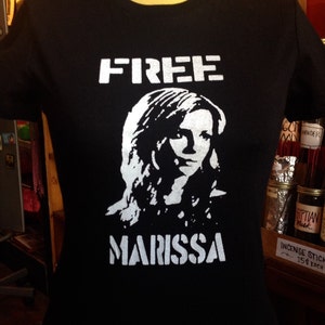 May include: Black t-shirt with white text that reads "FREE MARISSA". The shirt features a white silhouette of a woman with long hair.