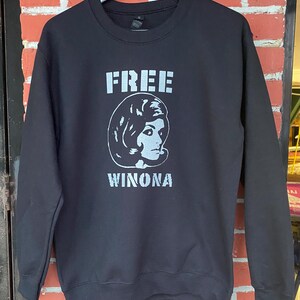 May include: Black sweatshirt with the text "FREE WINONA" in a light grey font. The design includes a stylized image of a woman's face. The sweatshirt is hanging on a black hanger against a brick wall.